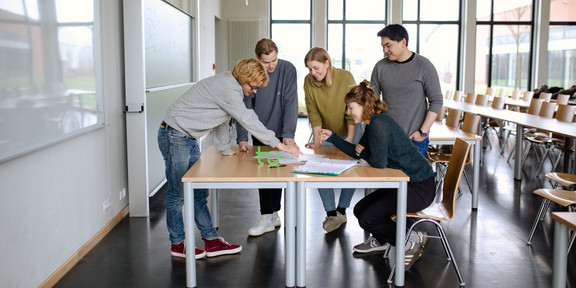 Studierende stehen gemeinsam um einen Tisch im Seminarraum und arbeiten.