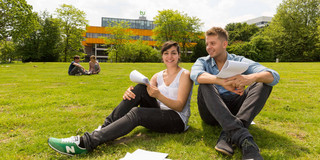 Zwei Studierende sitzen auf einer Wiese, im Hintergrund die Mensa