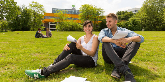 Zwei Studierende sitzen auf einer Wiese, im Hintergrund die Mensa