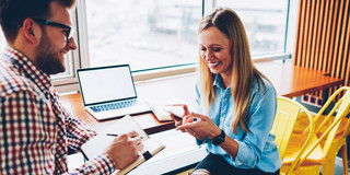 a man and a woman are sitting at a desk facing each other. On the desk is an open laptop