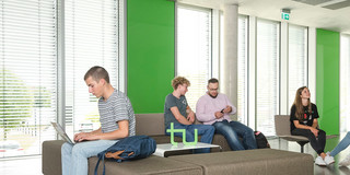 Six students are sitting at a table reading books, working with laptops or talking to each other
