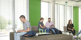 Six students are sitting at a table reading books, working with laptops or talking to each other