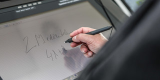 Bildschirm eines Hörsaalpults, an dem jemand mit einem Stift in der Hand schreibt.
