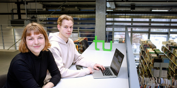 Zwei Studierende sitzen an einem langen Tisch vor einem Laptop in der Bibliothek.