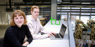 Zwei Studierende sitzen an einem langen Tisch vor einem Laptop in der Bibliothek.
