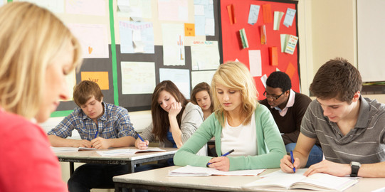 Schülerinnen und Schüler sitzen in einer Klasse und schreiben in Hefte, vor ihnen sitzt eine Lehrerin an einem Pult.
