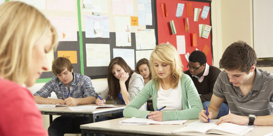 Pupils sit in a class and write in notebooks sitting in front of them a teacher at a desk.