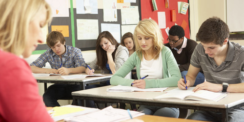Schülerinnen und Schüler sitzen in einer Klasse und schreiben in Hefte. Vor ihnen sitzt eine Lehrerin an einem Pult.