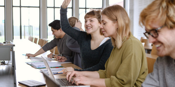 Studierende im Seminarraum sitzen in einer Tischreihe vor ihren Laptops. 