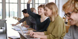 Five students sit in the seminar room with their laptops, one of them give a hand signal.