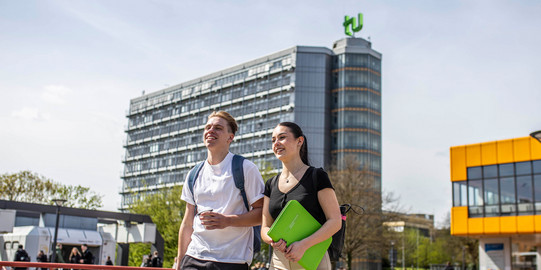 Ein junger Mann und eine junge Frau laufen über die Mensabrücke. Im Hintergrund sieht man den Mathetower mit TU-Logo und das Mensagebäude.