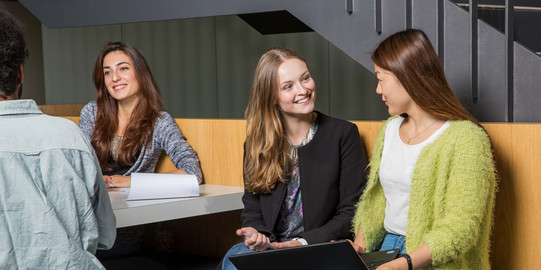 Zwei Studierende unterhalten sich und sitzen gemeinsam auf einer Bank mit einem Laptop.