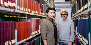 Two students stand between two bookshelves in the library.