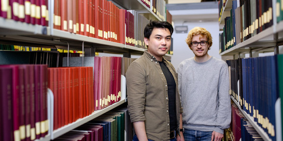 Two students stand between two bookshelves in the library.