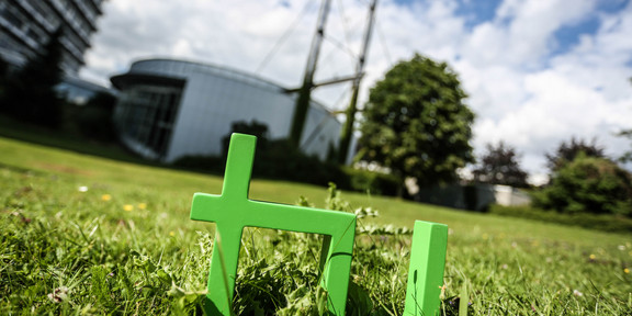 TU-Logo Im Vordergrund steht das TU-Logo im Gras, im Hintergrund das Audimax.