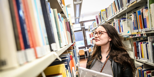 Studierende steht mit einem Tablet in der linken Hand an den Bücherregalen in der Bibliothek und sucht ein Buch.