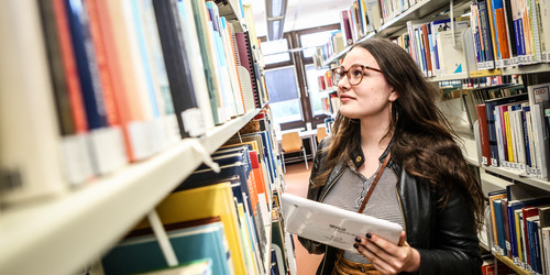 A student holding a tablet in her left hand is searching for a book in the library.
