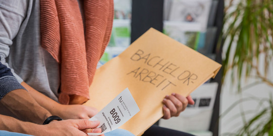 Bachelorarbeit Zwei Personen sitzen nebeneinander. Man sieht einen Ausschnitt von Knie bis Brust. Die hintere Person hat einen großen Umschlag mit der Aufschrift „Bachelor-Arbeit“ in der Hand, die vordere Person hat einen kleinen weißen Zettel mit einer Wartenummer, einem Code und dem TU-Logo in der Hand.