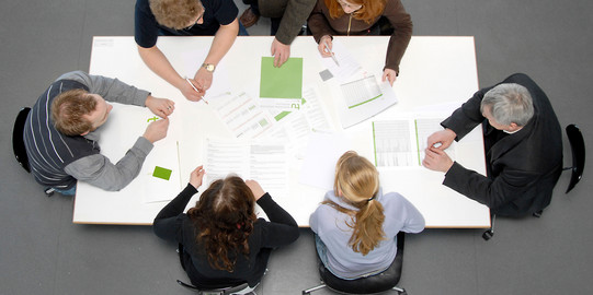 Studierende und Dozierende sitzen gemeinsam an einem Tisch und arbeiten.