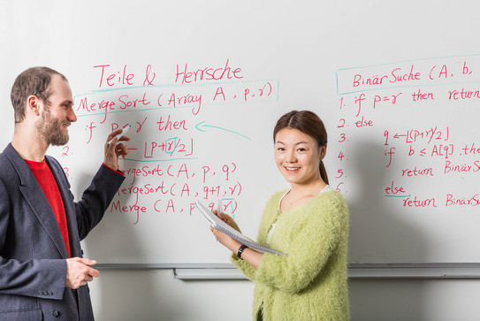 Zwei Personen stehen vor einem Whiteboard mit Formeln und Beschriftung. Eine Person hat einen Stift in der Hand, die andere einen Block.