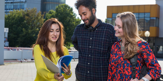 Three students are standing together in a square in front of two buildings.