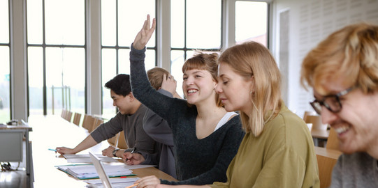 Five students sitting in the seminar room at the table. A female student raises her hand.