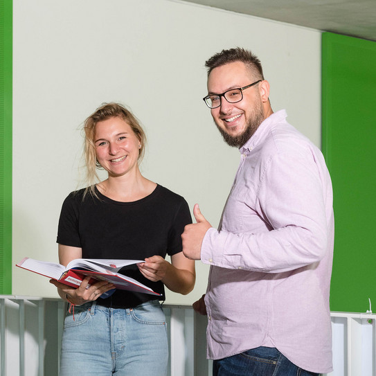 Studierende vor Geländer, Daumen hoch Zwei Studierende stehen vor einem Geländer, einer streckt den Daumen hoch.