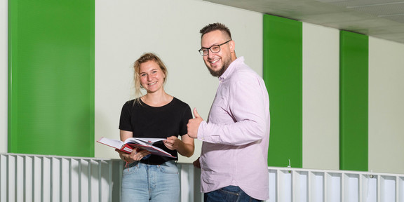 Zwei Studierende stehen vor einem Geländer, einer streckt den Daumen hoch.