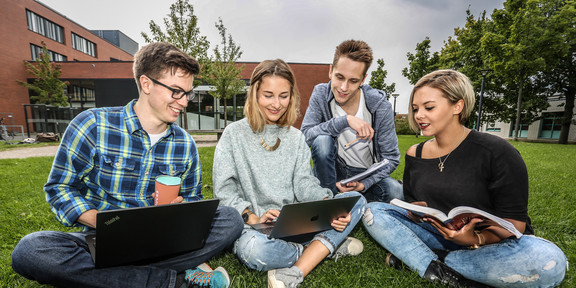 Four students are sitting on the grass and looking in a book.